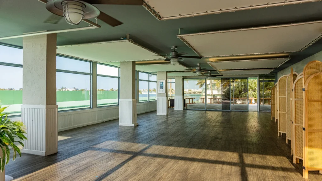 A spacious, empty room with large windows, ceiling fans, and wooden flooring. Sunlight streams in, highlighting the bright, airy space. There are folding screens on one side and a view of palm trees outside.