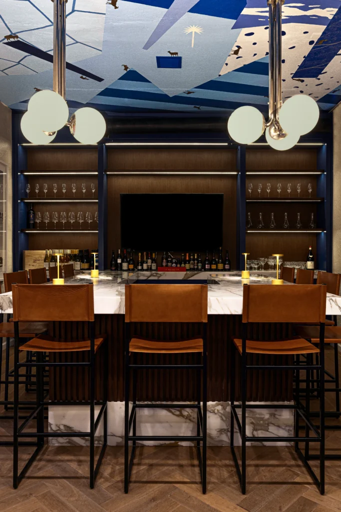 A modern bar with a marble countertop, tan leather barstools, a wall of shelves holding glassware and wine bottles, globe pendant lights, and a blue geometric ceiling design.