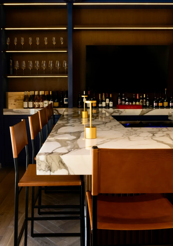 A modern bar with a marble countertop, brown leather bar stools, gold accents, shelves of glassware and liquor bottles, and a large TV mounted on the wall behind the bar.