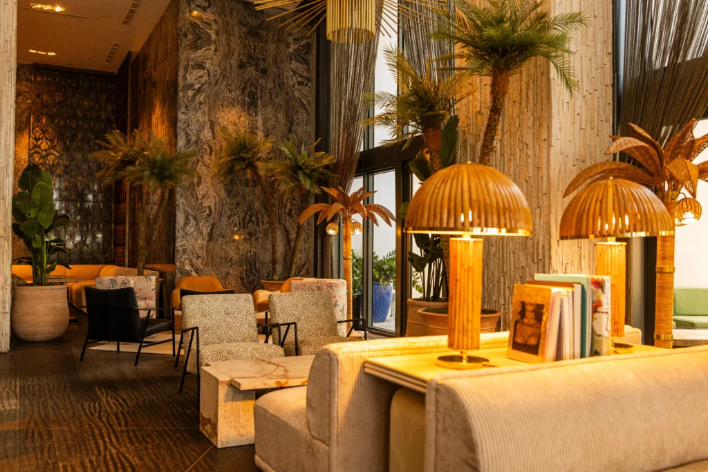 A stylish hotel lounge with cozy seating, marble walls, indoor plants, and warm lighting from rattan lamps on a table, creating a welcoming atmosphere.