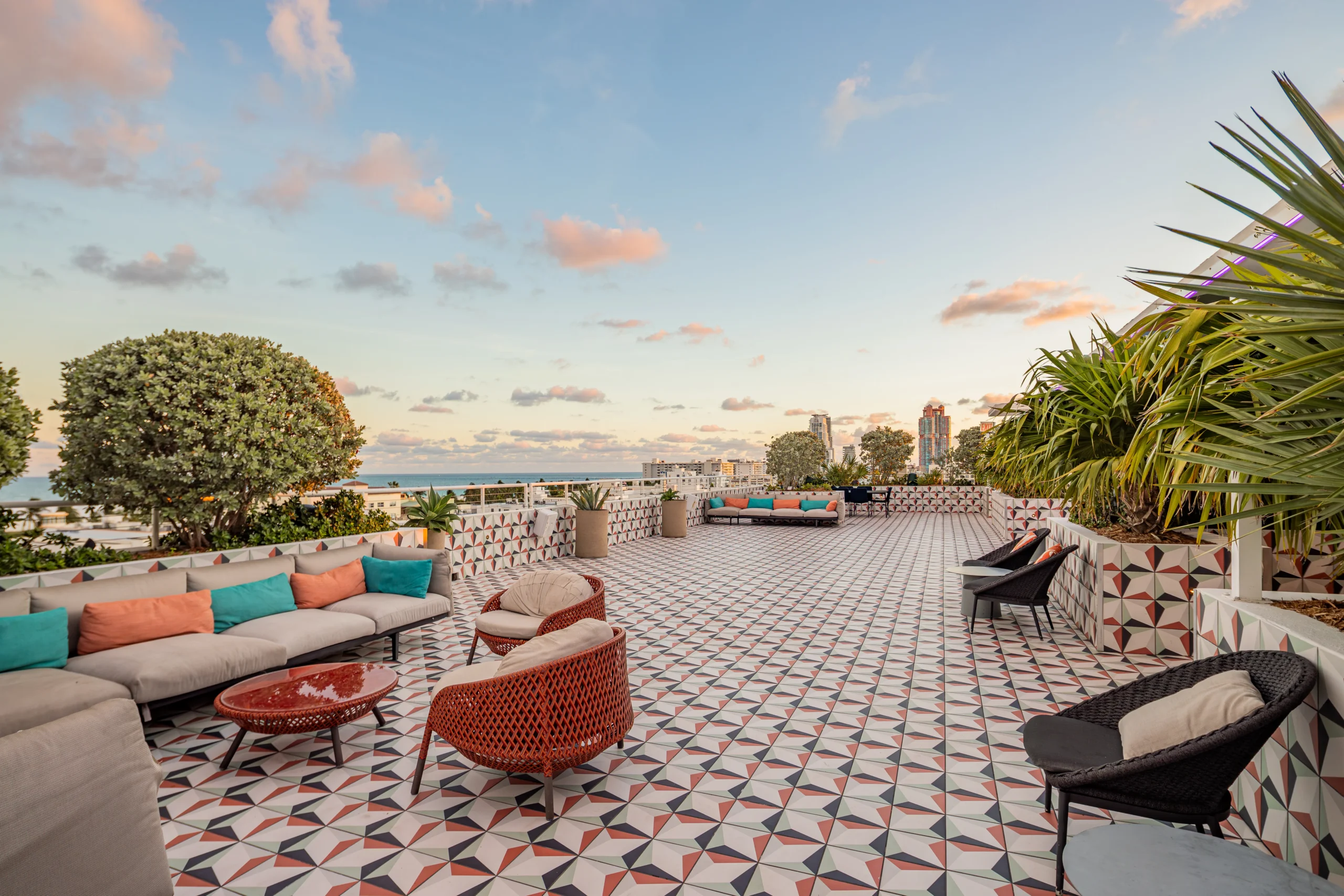 A spacious rooftop terrace with patterned tile flooring, modern outdoor seating, lush green plants, and a view of the city skyline and ocean under a partly cloudy sky at sunset.