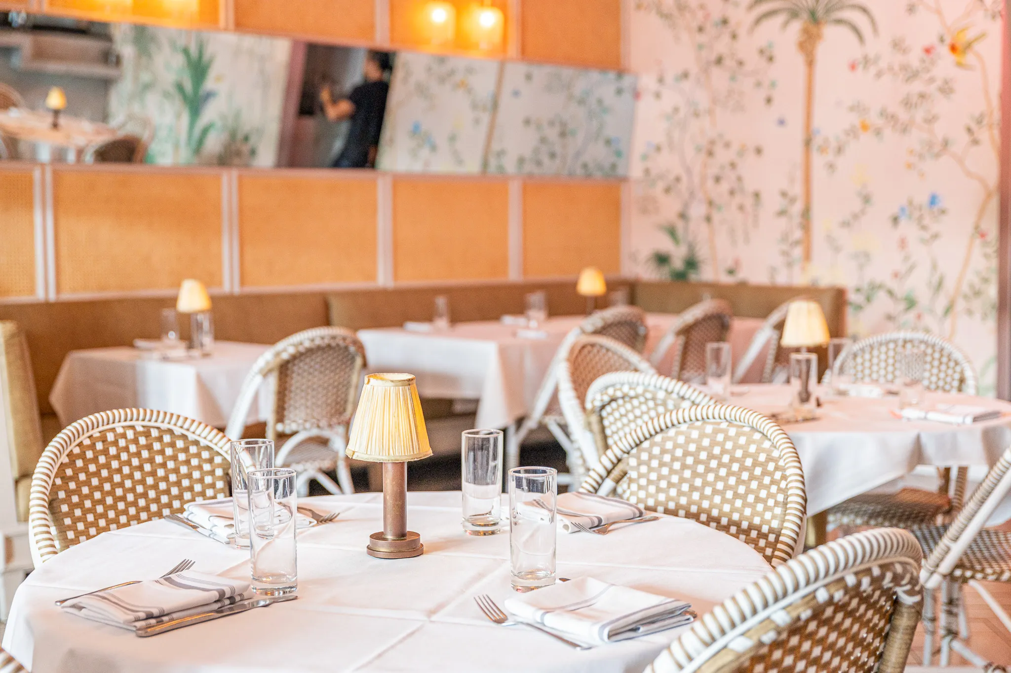 A bright, elegant restaurant dining area with white tablecloths, wicker chairs, small table lamps, glassware, and napkins. The walls feature pastel floral and palm tree wallpaper, creating a cozy, inviting atmosphere.