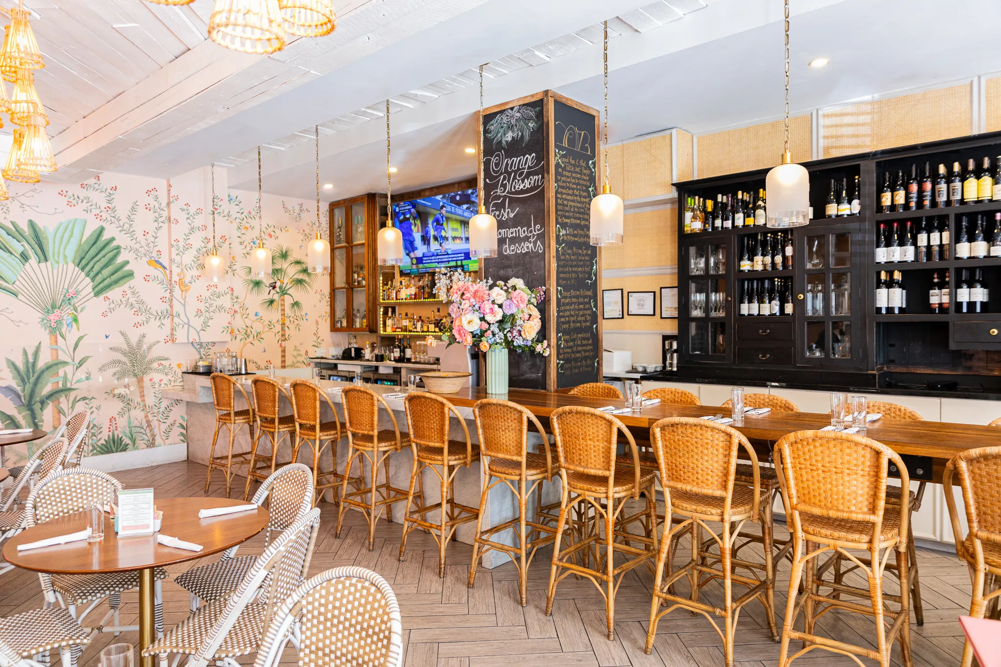 A bright, airy restaurant bar with wicker barstools, floral wallpaper, hanging pendant lights, a chalkboard menu, neatly arranged wine bottles, and fresh flowers on the bar counter. Round tables are set nearby.
