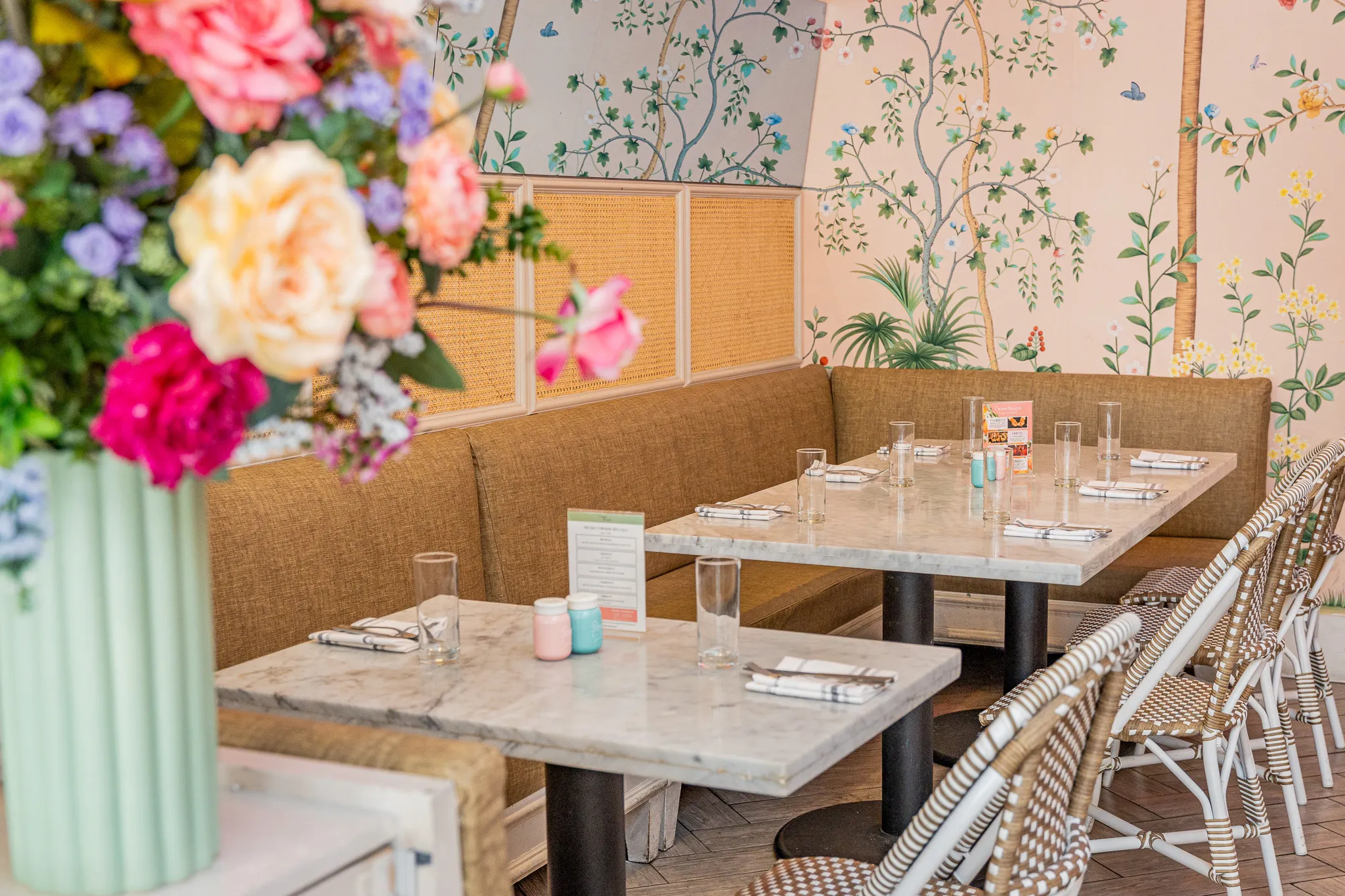 A cozy restaurant features marble tables set with glasses and napkins, wicker chairs, a tan cushioned bench, and floral wallpaper. A large vase of colorful flowers is in the foreground on the left.