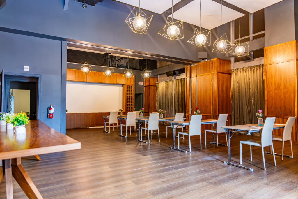A spacious, modern meeting room with wooden floors, several tables and chairs arranged in rows, pendant lights hanging from the ceiling, and large windows with curtains along one side.