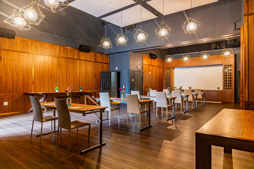 A modern conference room with wooden walls, stylish pendant lights, tables spaced apart with white chairs, and a projector screen at the front. Small vases with flowers are on the tables. The room has a polished wooden floor.