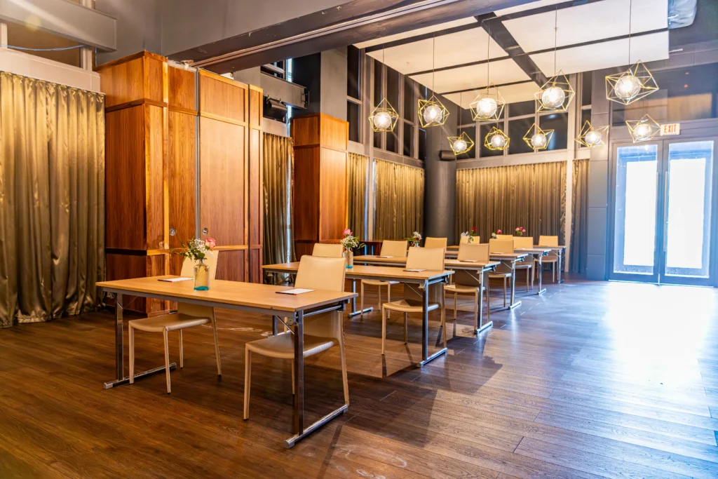 A modern conference room with wooden floors, several tables and chairs arranged in rows, each table with a notebook, pen, and vase of flowers. Geometric pendant lights hang from the ceiling, and curtains cover the windows.