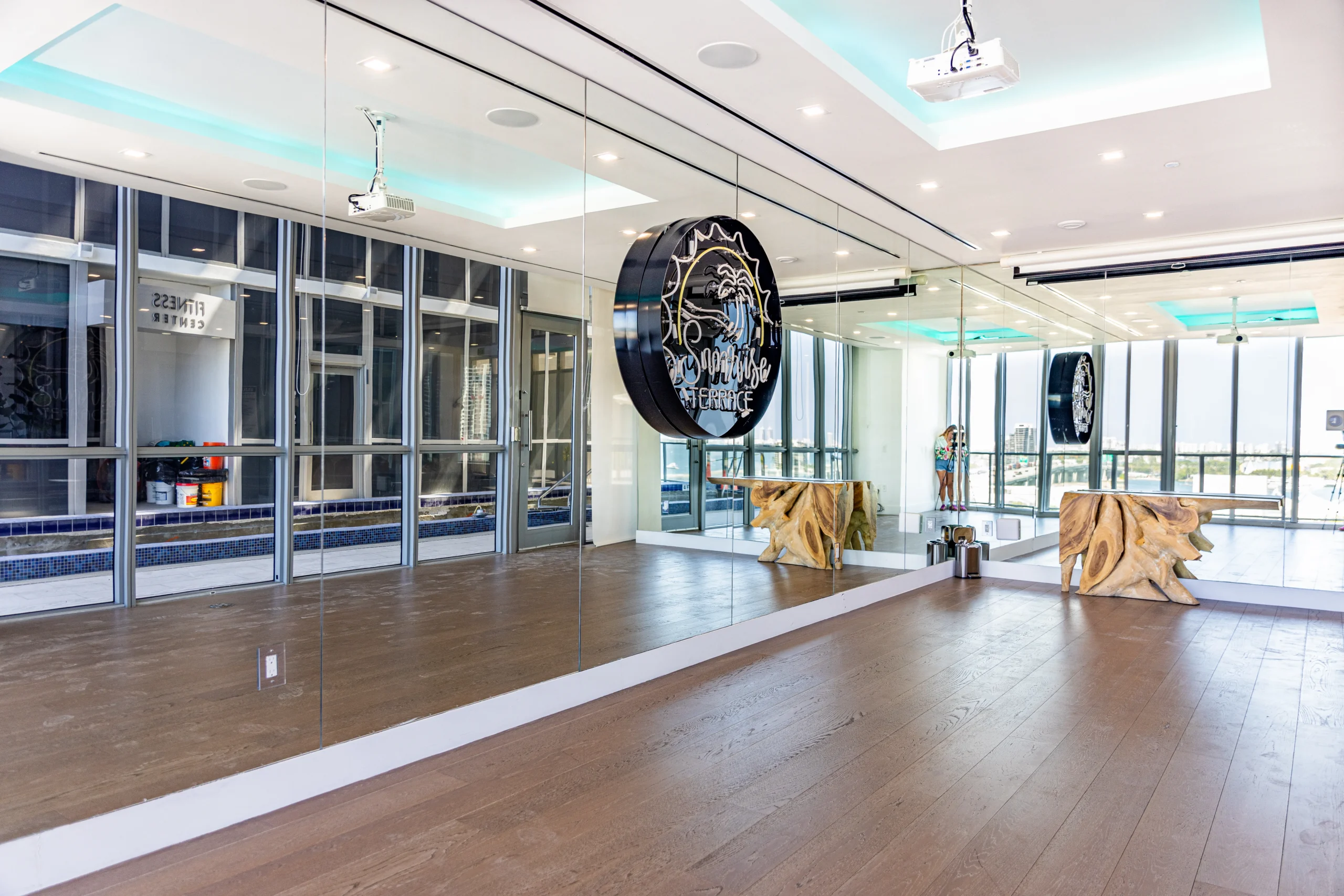 A modern fitness studio with wood flooring, mirrored walls, a decorative wooden table, and a large circular sign. Floor-to-ceiling windows reveal a pool and city view outside. Bright, recessed lighting illuminates the space.