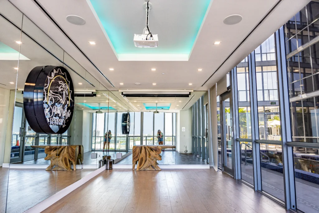 Modern fitness studio with wood floors, mirrored walls, a neon sign reading “Dogpound Terrace,” decorative golden dog statues, yoga mats, and large windows showing an outdoor terrace and city view.