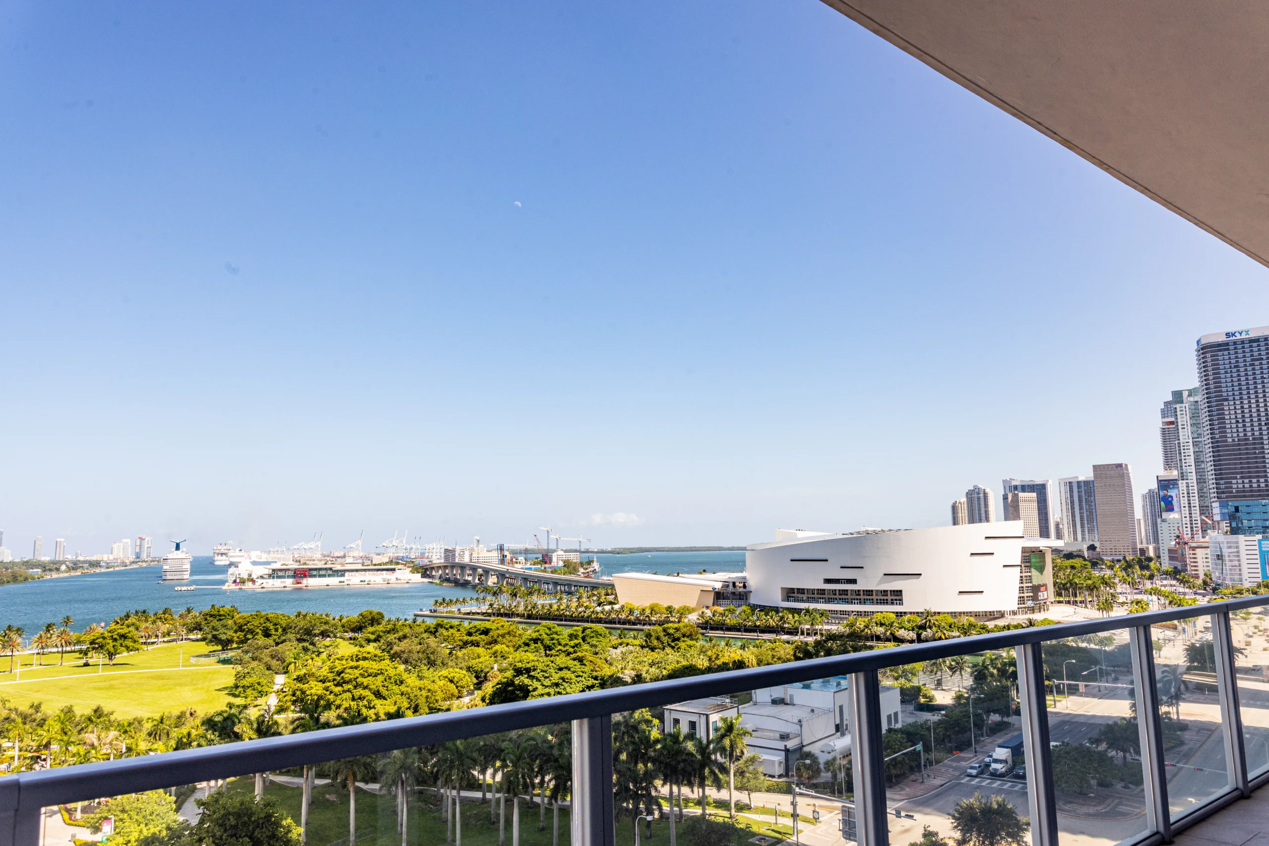 View from a balcony overlooking a cityscape with tall buildings, a modern white structure, green trees, a harbor with ships, and a blue sky with the ocean in the background.