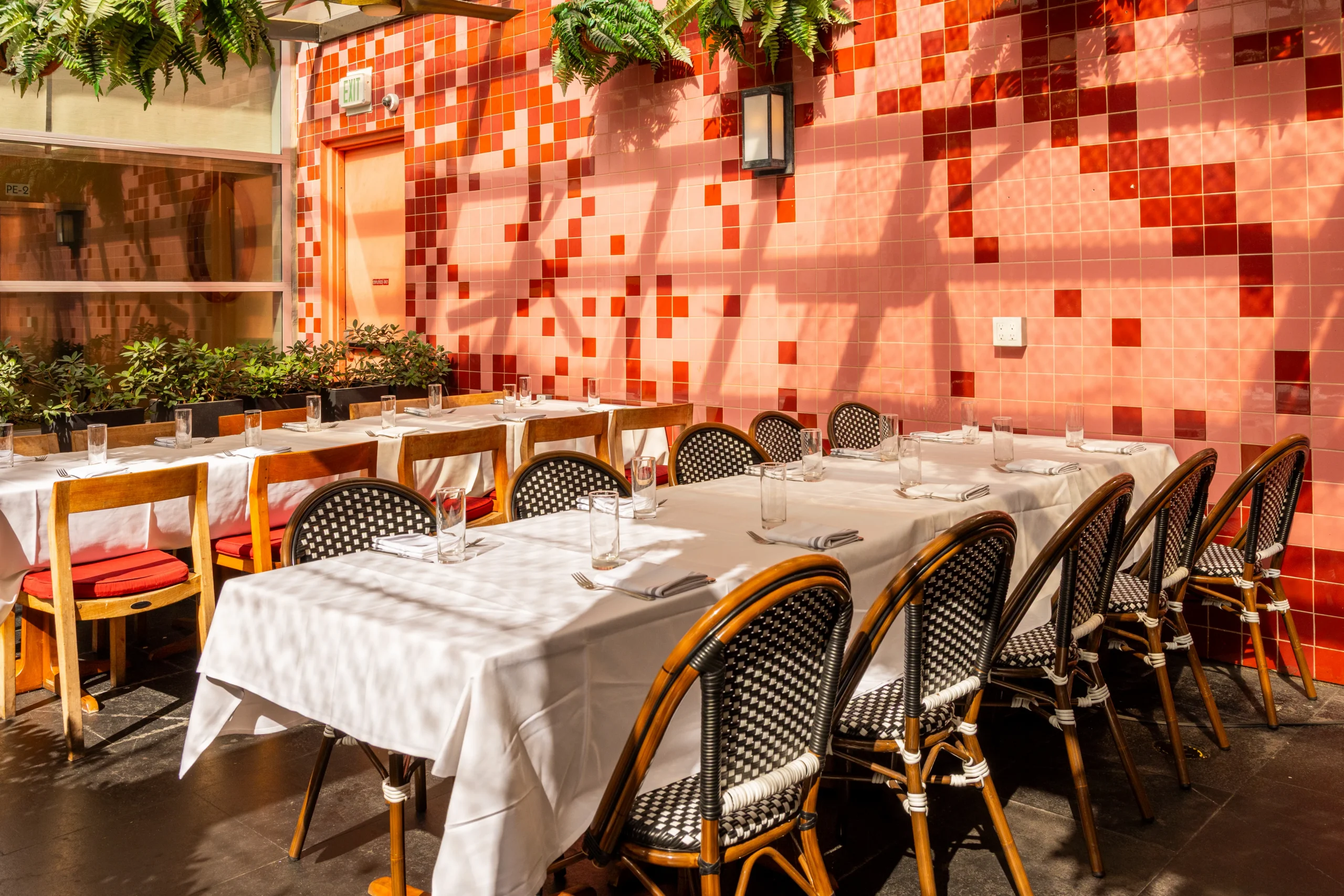 A sunlit restaurant dining area with two long tables covered in white cloths, set with glasses and napkins, surrounded by wicker chairs. The pink and red tiled wall and hanging plants create a bright, inviting atmosphere.