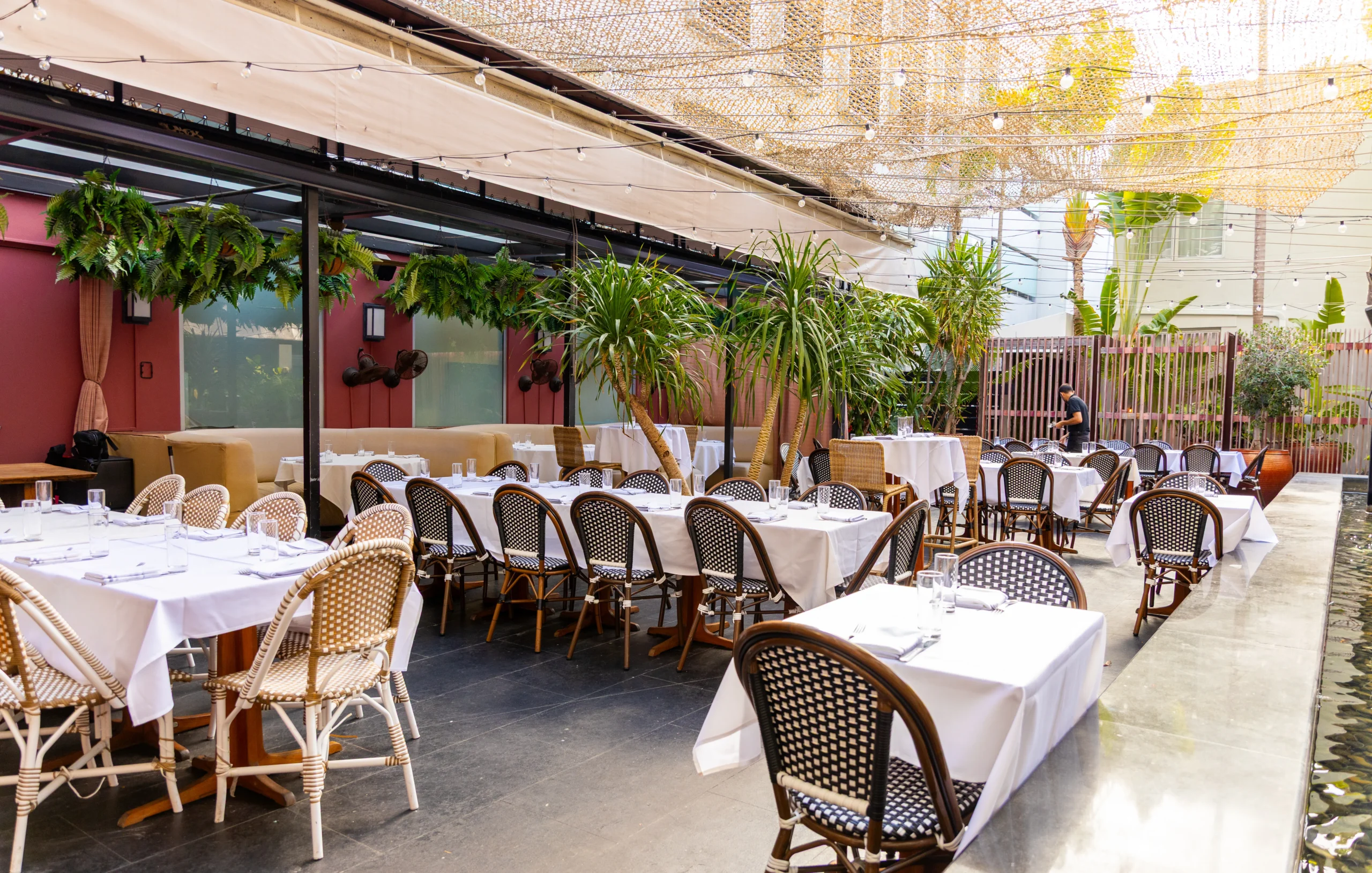 An outdoor restaurant patio with tables covered in white cloths, wicker chairs, potted plants, and hanging greenery. String lights and a mesh canopy provide shade, creating a welcoming, elegant dining atmosphere.