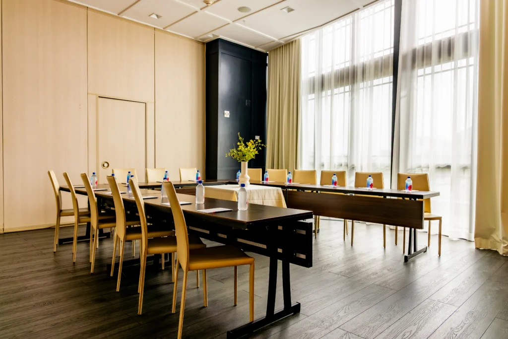 A modern conference room with two tables arranged in a U-shape, surrounded by wooden chairs. Each place setting has a water bottle and notepad. Large windows with sheer curtains let in natural light. A vase with flowers sits on the table.