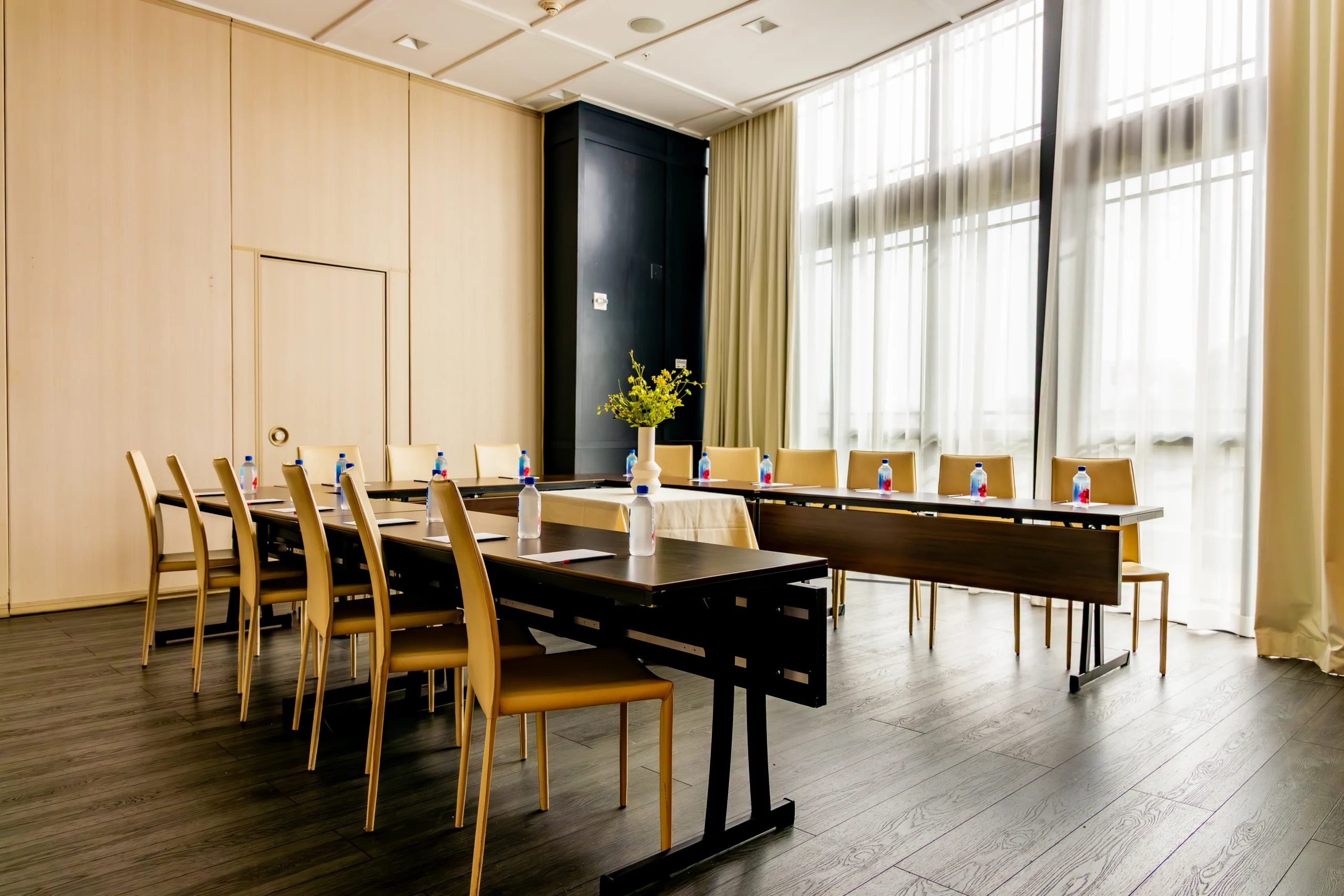A modern conference room with two tables arranged in a U-shape, surrounded by wooden chairs. Each place setting has a water bottle and notepad. Large windows with sheer curtains let in natural light. A vase with flowers sits on the table.