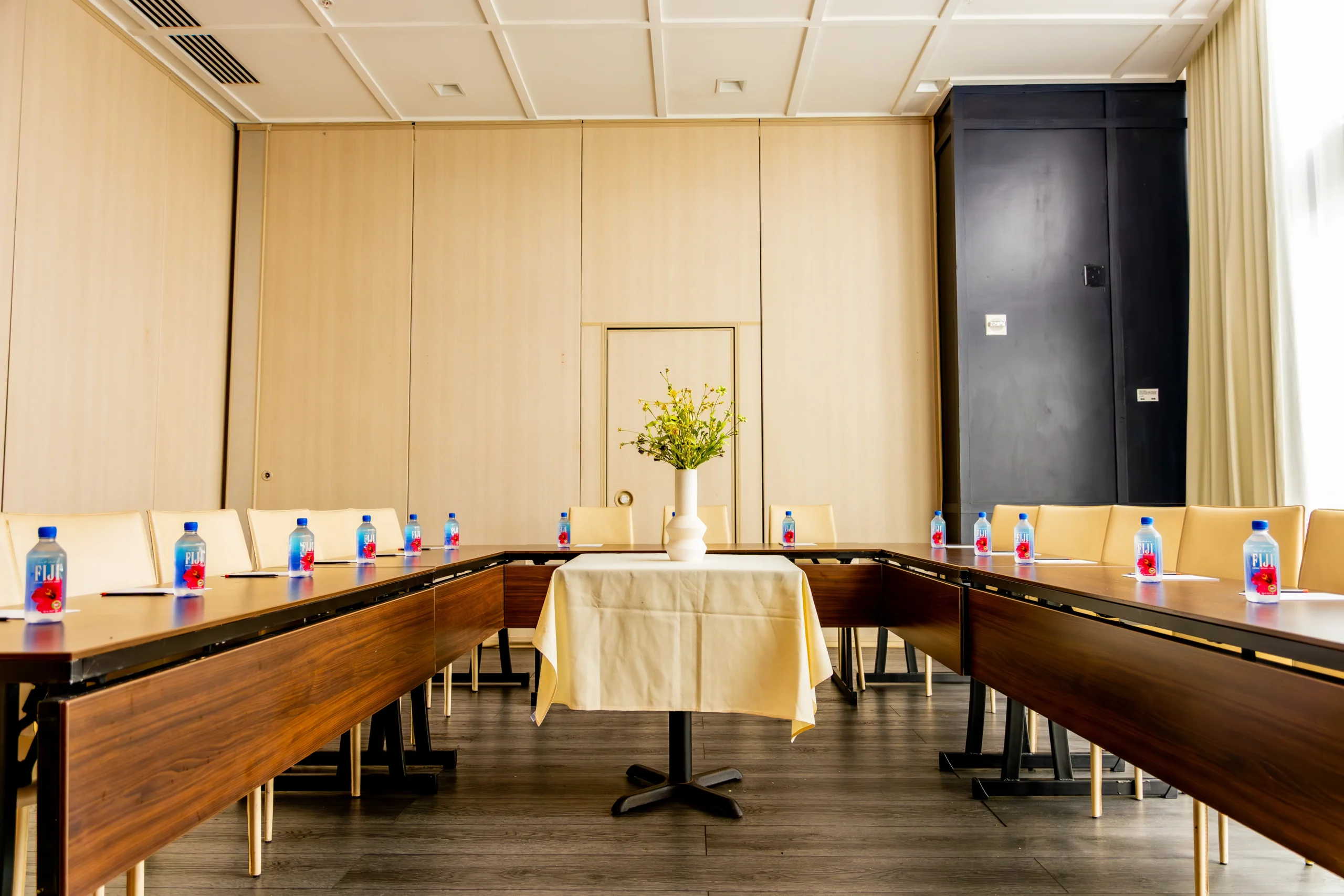 A conference room with U-shaped tables, each place set with a notepad, pen, and bottled water. A vase with yellow flowers is on a small table in the center of the room.