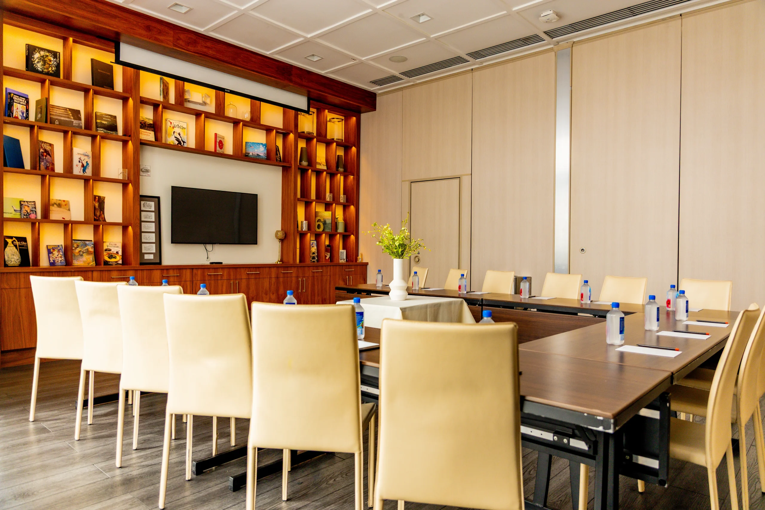 A modern conference room with a U-shaped table, cream chairs, water bottles, notepads, and a vase of flowers. Shelves with books and decorations line the back wall, and a flat-screen TV is mounted near the center.