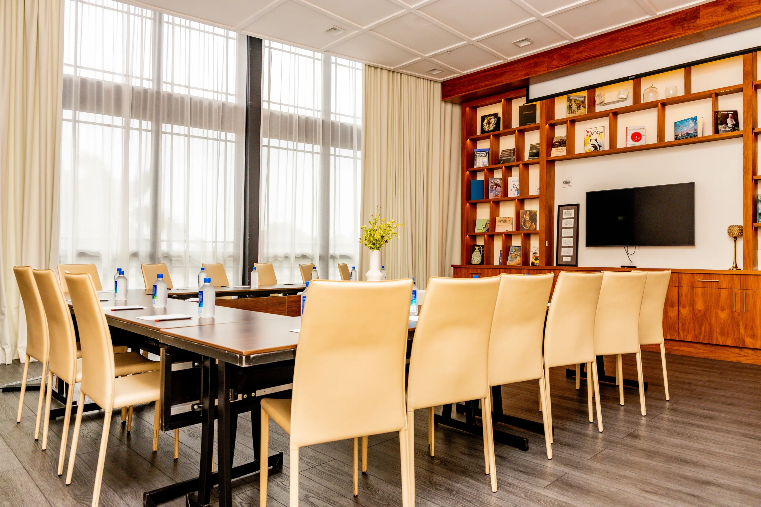 A modern conference room with a large table surrounded by beige chairs, water bottles on the table, a TV on the wall, shelves with books and decorations, and tall windows with sheer curtains letting in natural light.