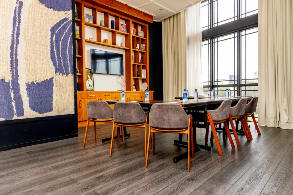 A modern conference room with a long table, eight gray chairs, water bottles, a large wall-mounted TV, wooden shelves with decor, floor-to-ceiling windows, and light curtains.
