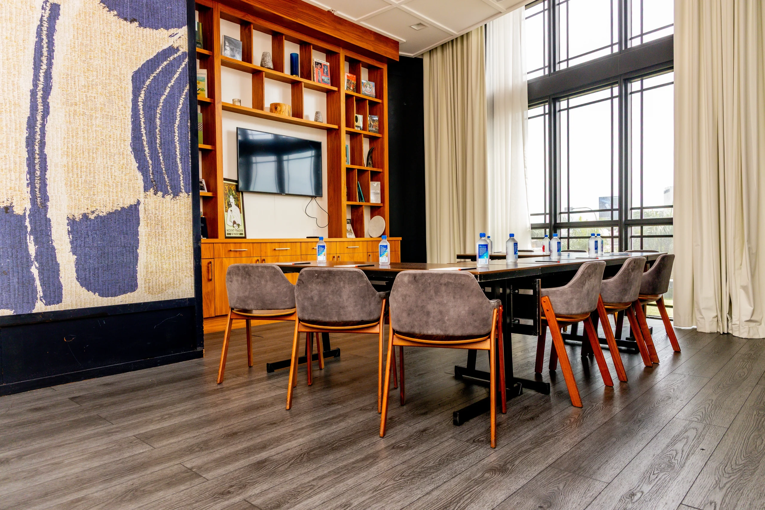 A modern conference room with a long table, eight gray chairs, water bottles, a large wall-mounted TV, wooden shelves with decor, floor-to-ceiling windows, and light curtains.