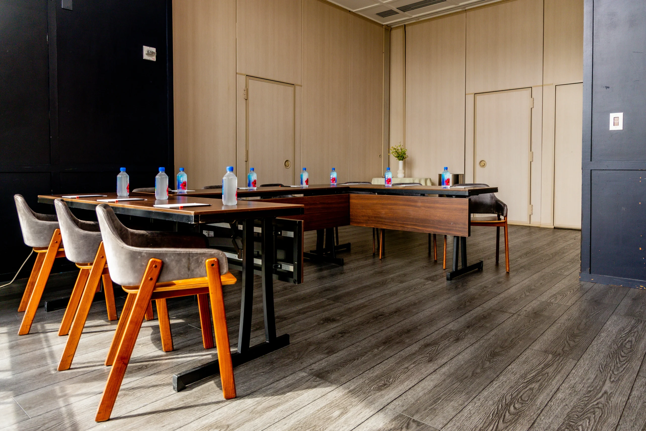 A modern, empty meeting room with wooden floors, several chairs around U-shaped tables, and bottled water placed at each seat. The room has light-colored walls and a small potted plant in the corner.
