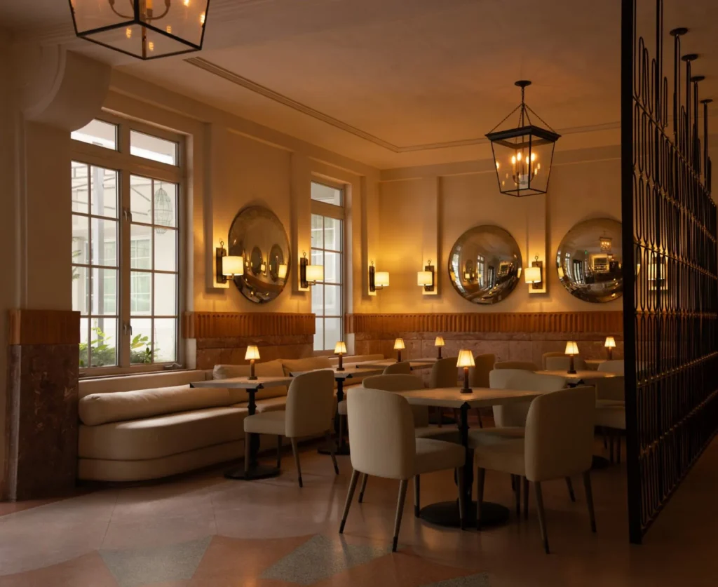 A cozy, elegant restaurant interior with cream-colored chairs, round tables with small lamps, large round mirrors on the walls, and warm lighting from ceiling lanterns and wall sconces. Large windows let in natural light.