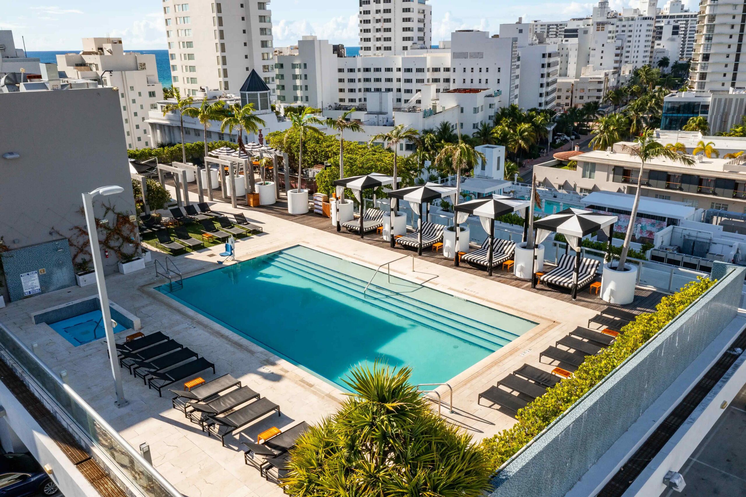 A rooftop swimming pool surrounded by lounge chairs and cabanas, with tall city buildings and palm trees in the background under a partly cloudy sky.