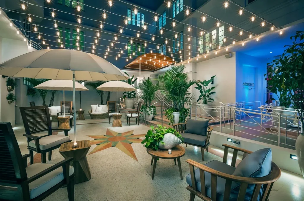 A stylish outdoor patio features string lights overhead, potted plants, cushioned wooden chairs, umbrellas, and small tables, creating a cozy, inviting atmosphere with a star design on the floor.