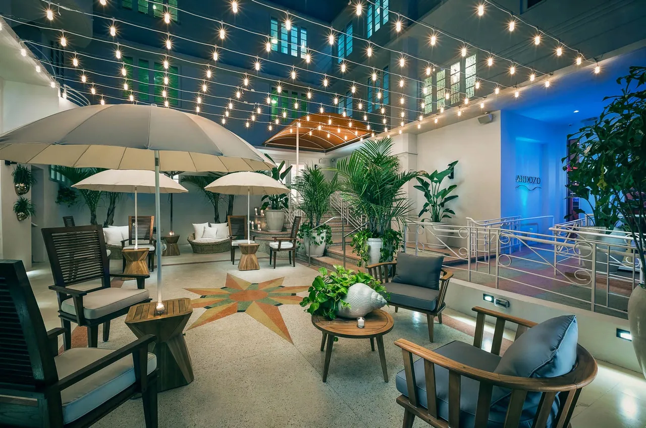 A stylish outdoor patio features string lights overhead, potted plants, cushioned wooden chairs, umbrellas, and small tables, creating a cozy, inviting atmosphere with a star design on the floor.