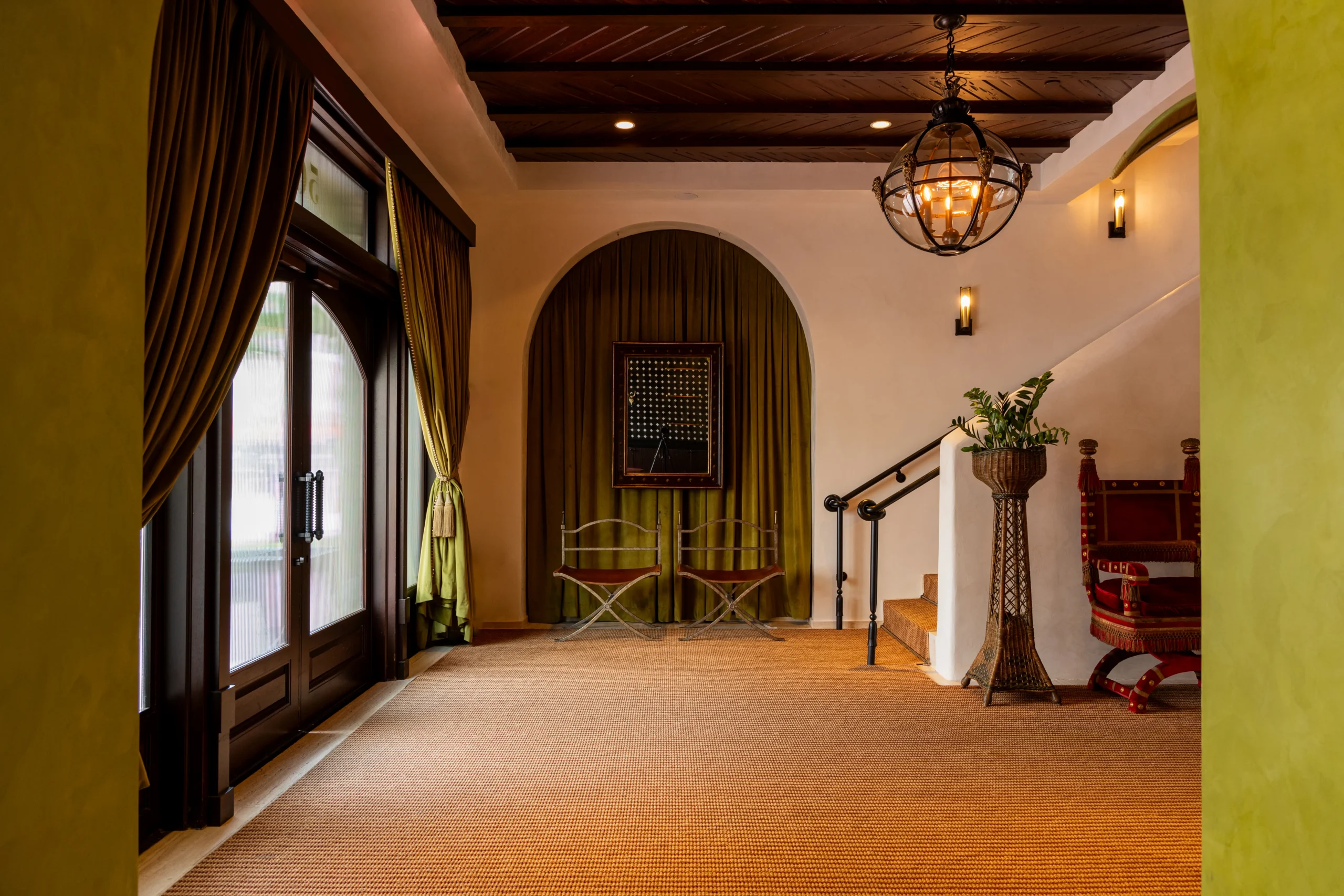 A warmly lit hallway with green curtains, a large glass double door, arched niches, two metal chairs, a red ornate chair, potted plant on the stair landing, and a classic lantern-style ceiling light.
