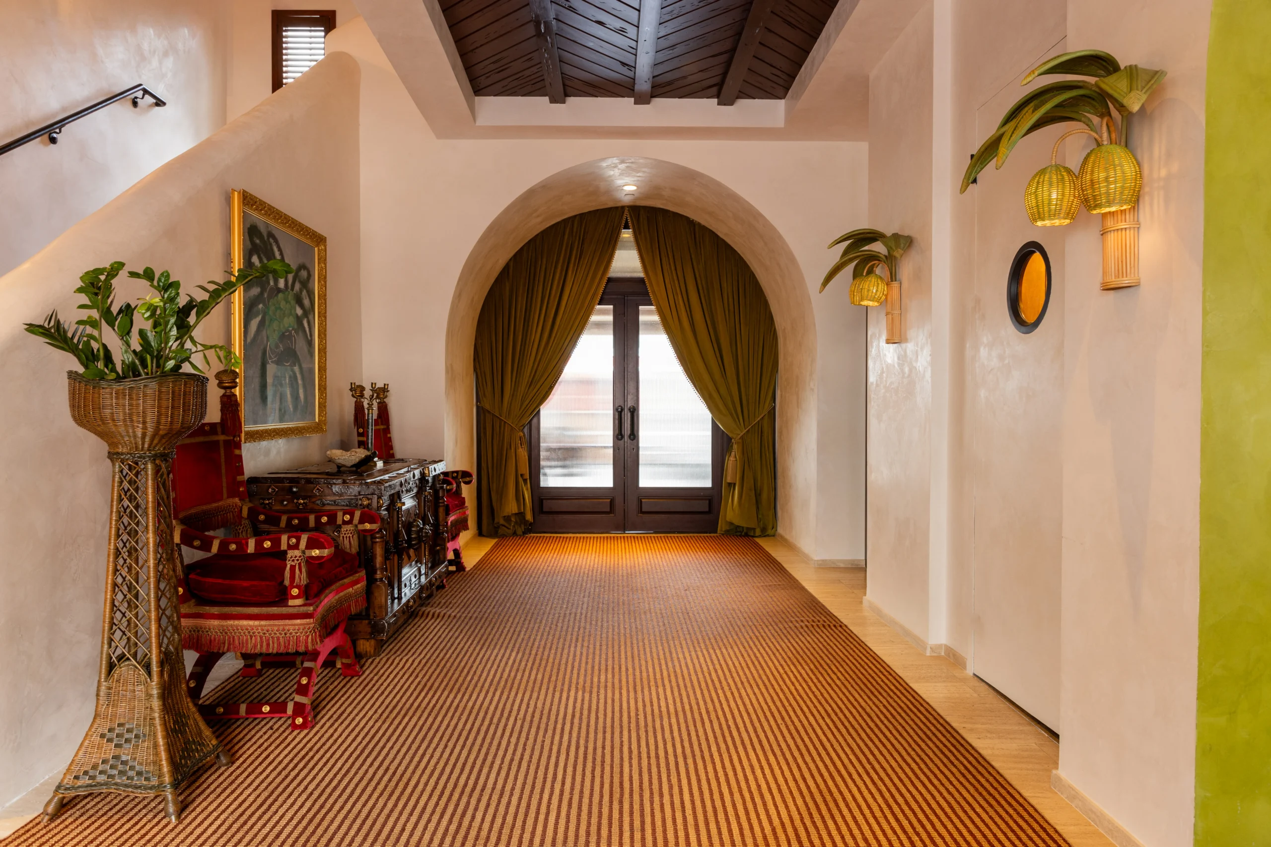 Elegant hallway with arched doorway and green curtains, woven rug, ornate wooden console, red upholstered chairs, potted plants, hanging wicker lights, and a large framed painting on a white wall.