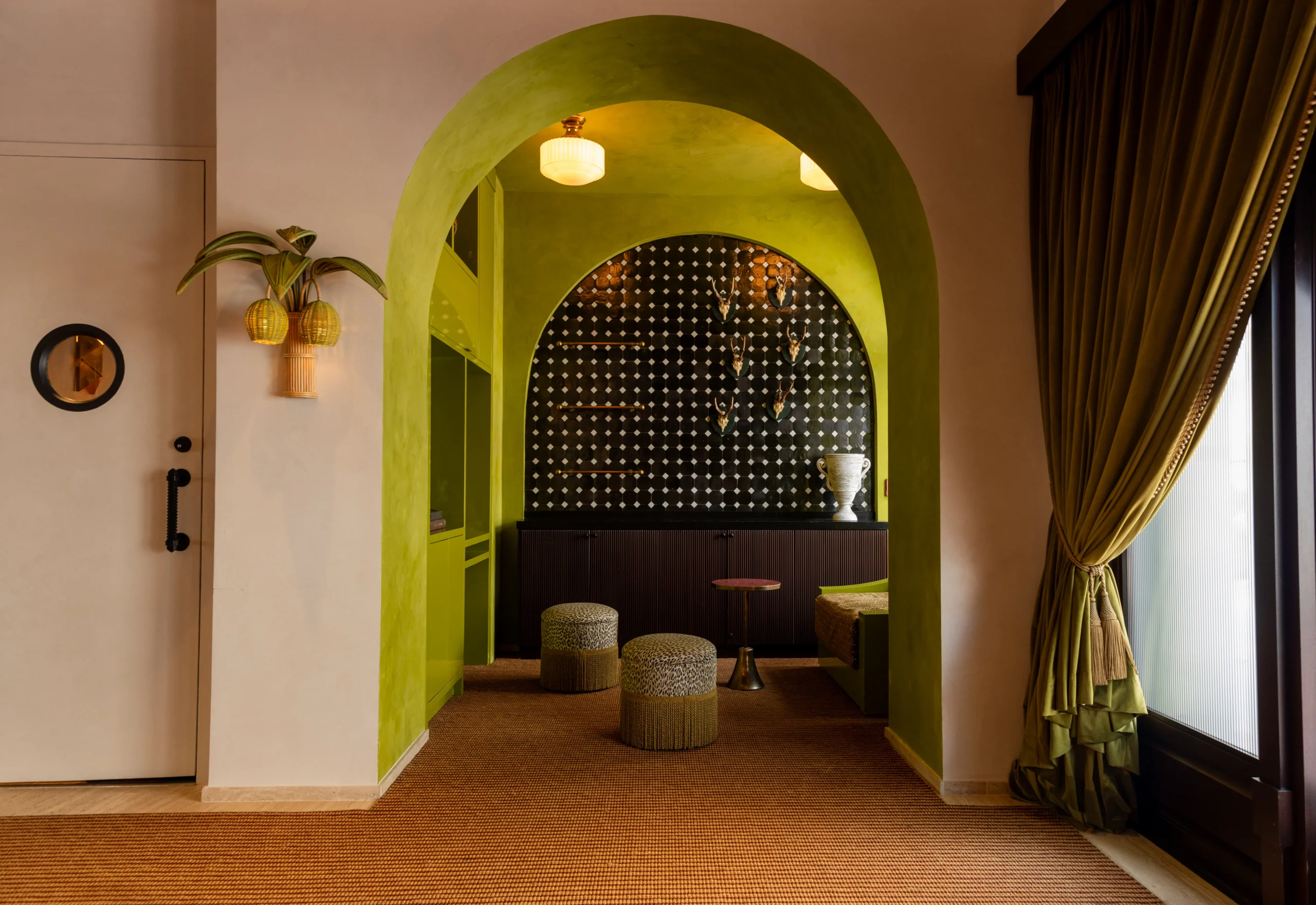 A stylish room with green archways, patterned wall, small poufs, a bench, gold pineapple wall sconce, potted plant, and floor-length green curtains next to a large window with soft natural light.