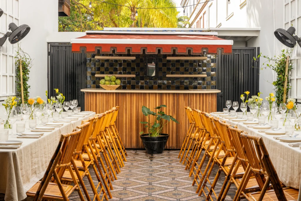 Long wooden tables with yellow flowers, set with glassware and plates, are lined with bamboo chairs facing a small bar with black tiles. The outdoor space is bright and inviting, surrounded by plants.