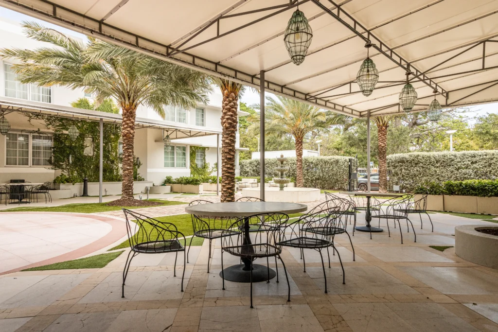 Outdoor patio with round tables and black metal chairs under a white canopy, surrounded by palm trees and greenery, with a modern building in the background. The area is spacious and inviting.