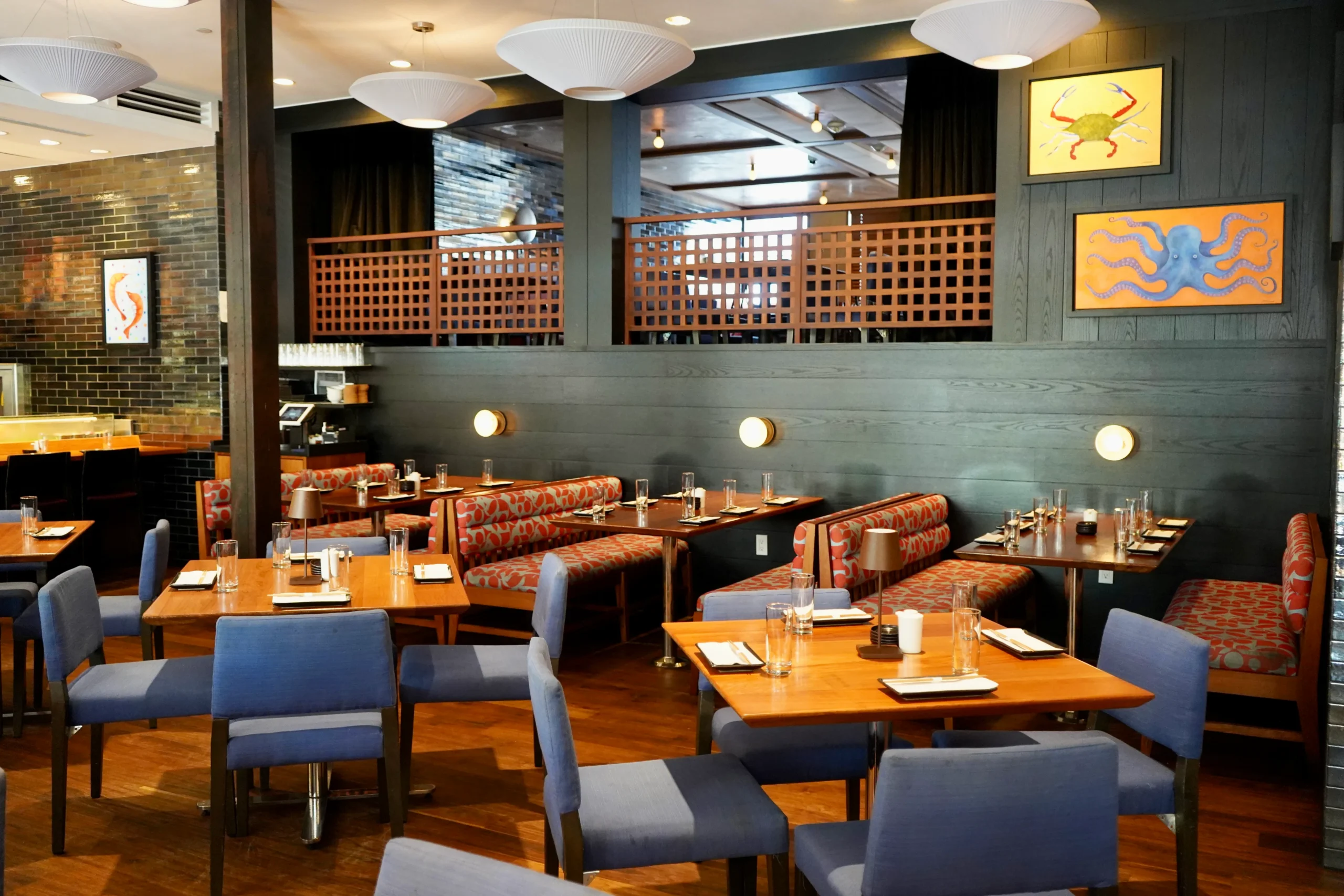 Modern restaurant interior with wooden tables, blue cushioned chairs, and patterned booth seating. Artworks with sea creatures hang on dark walls, and place settings are arranged neatly on each table.
