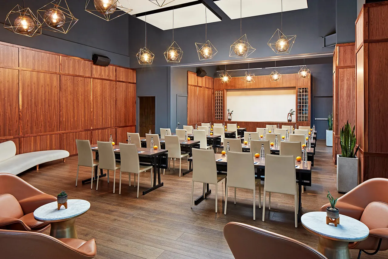 Modern conference room with wooden floors and walls, arranged with white chairs and black tables facing a large presentation screen. Warm hanging lights and minimalist decor, including plants and small tables, create a stylish atmosphere.