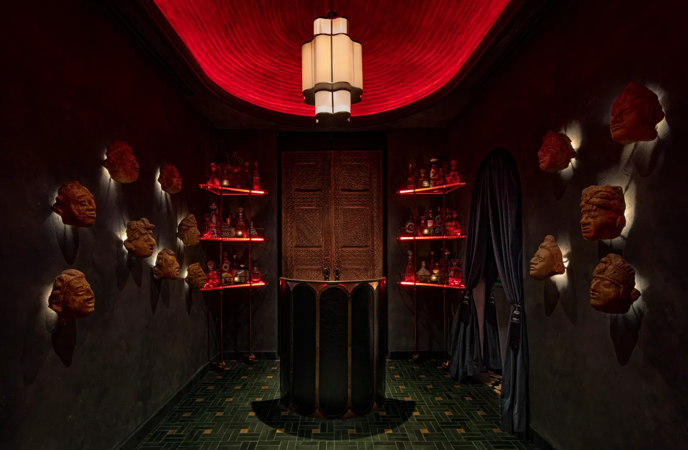 A dimly lit room with a red glowing ceiling, sculpted heads mounted on dark walls, shelves displaying bottles, a black counter in the center, and double wooden doors at the back. A draped object stands to the right.