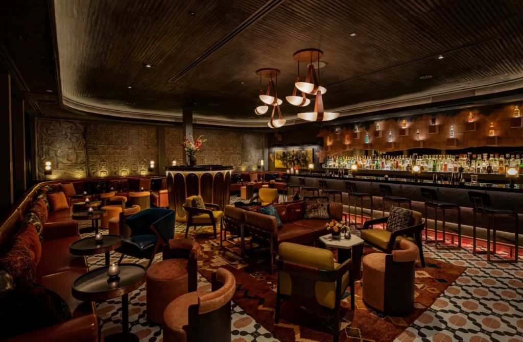 A stylish, dimly lit lounge bar with plush seating, patterned rugs, small round tables, and a long bar lined with bottles and stools. Warm lighting and rich decor create an inviting, elegant atmosphere.