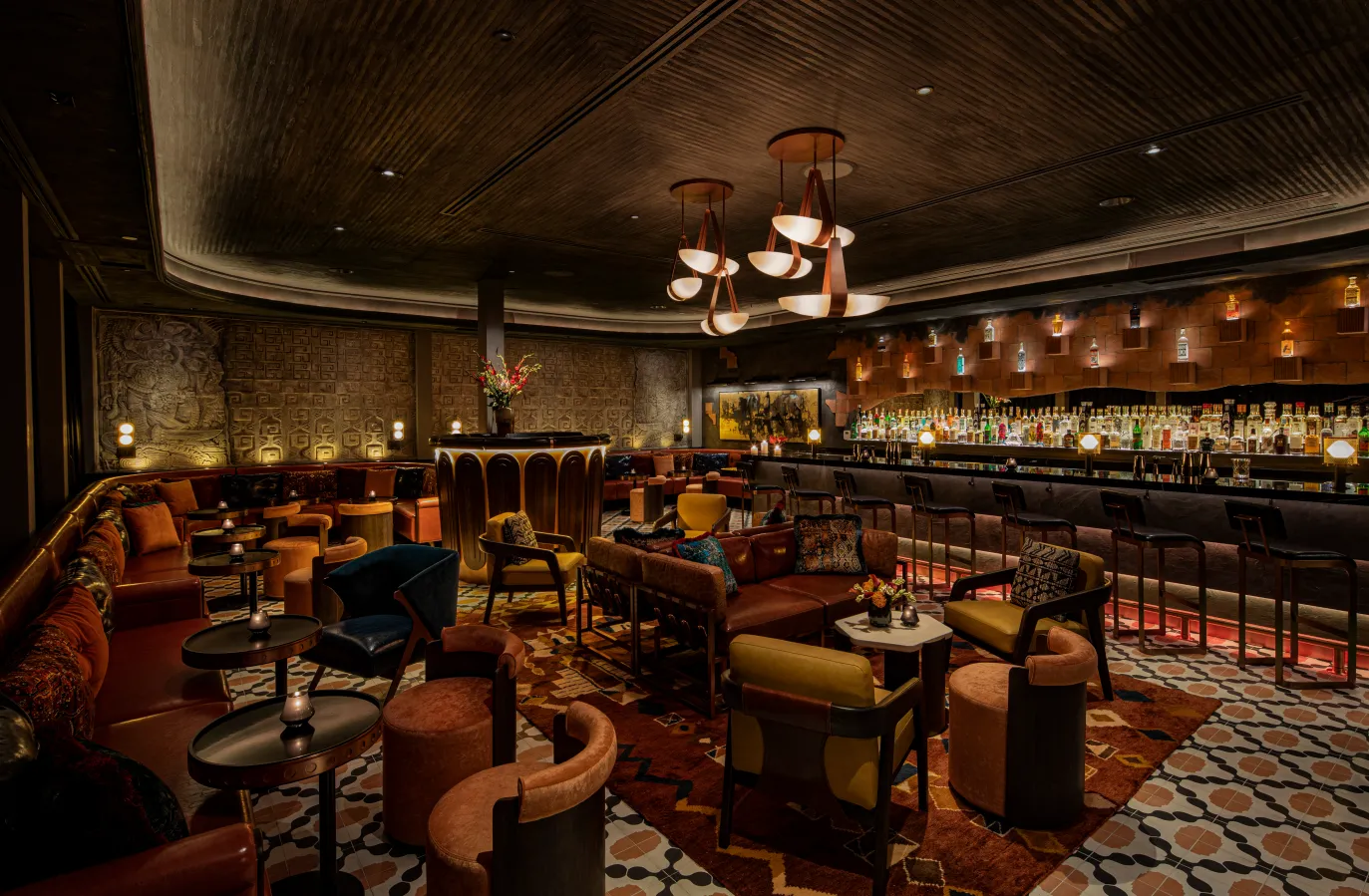 A stylish, dimly lit lounge bar with plush seating, patterned rugs, small round tables, and a long bar lined with bottles and stools. Warm lighting and rich decor create an inviting, elegant atmosphere.