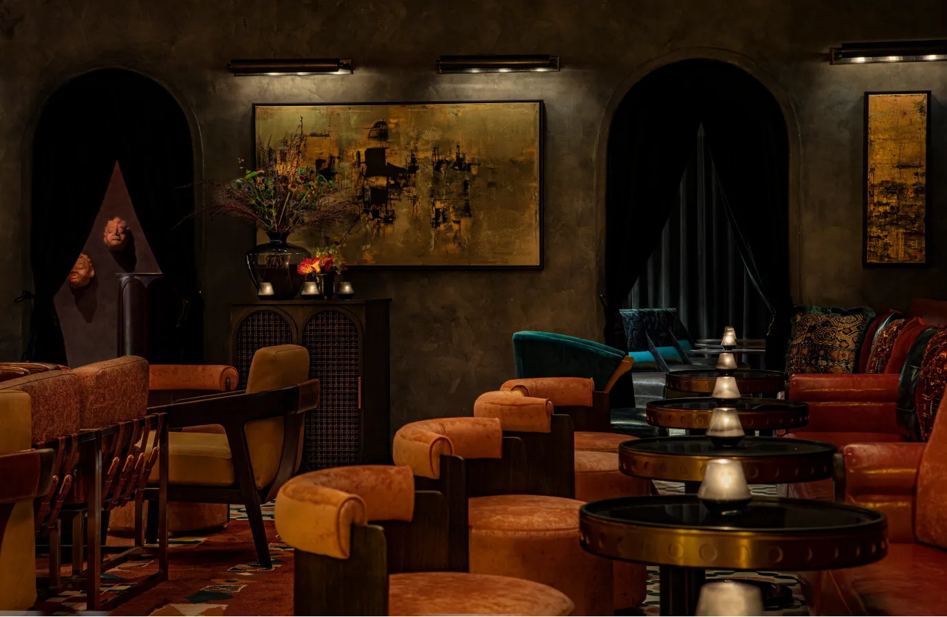 Dimly lit lounge with orange velvet chairs, round dark tables with candles, abstract art on dark walls, and a geometric sculpture. Archways lead to a cozy seating area with teal armchairs in the background.