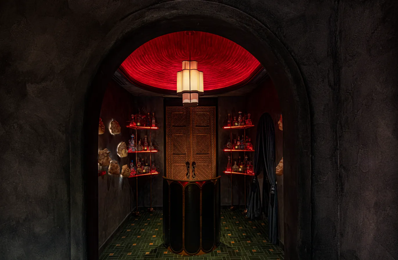 A dimly lit, arched room with dark walls, red lighting on the ceiling, shelves containing bottles and skulls, and a decorative wooden double door at the center beneath a modern hanging light.