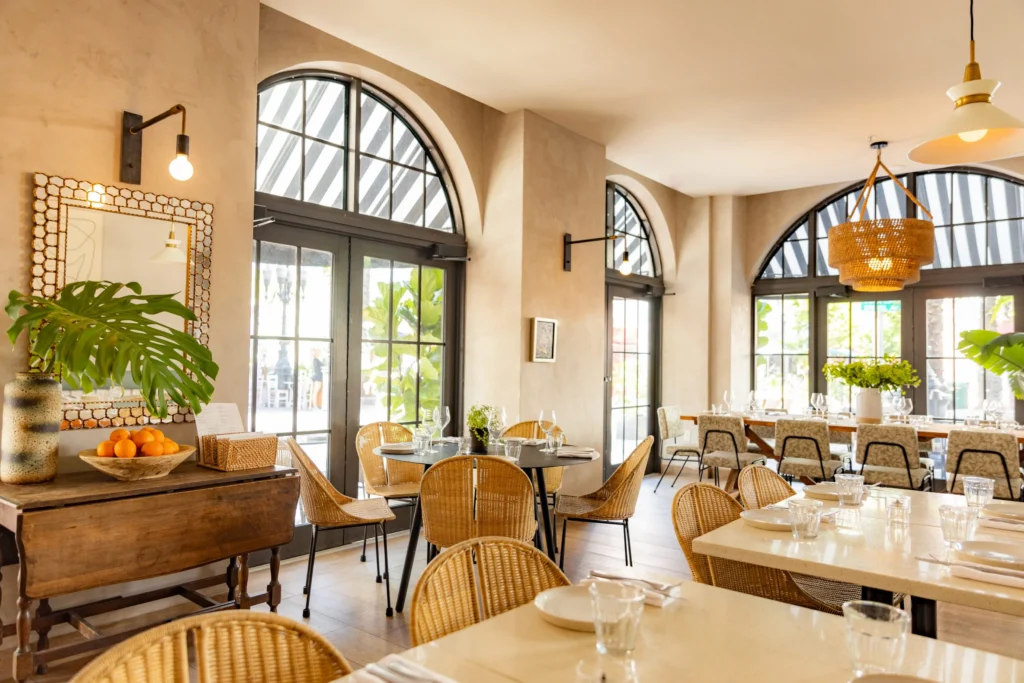 Bright, airy restaurant interior with large arched windows, rattan chairs, light wooden tables, and green plants. Sunlight streams in, creating a warm, inviting atmosphere with neatly set tables.