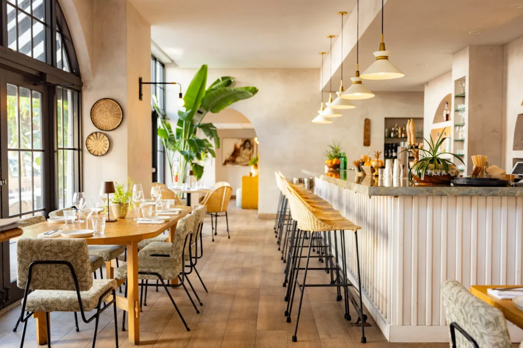 A bright, modern restaurant interior with wooden floors, a long bar with high stools, tables set for dining, large windows, potted plants, and pendant lights creating a cozy, inviting atmosphere.