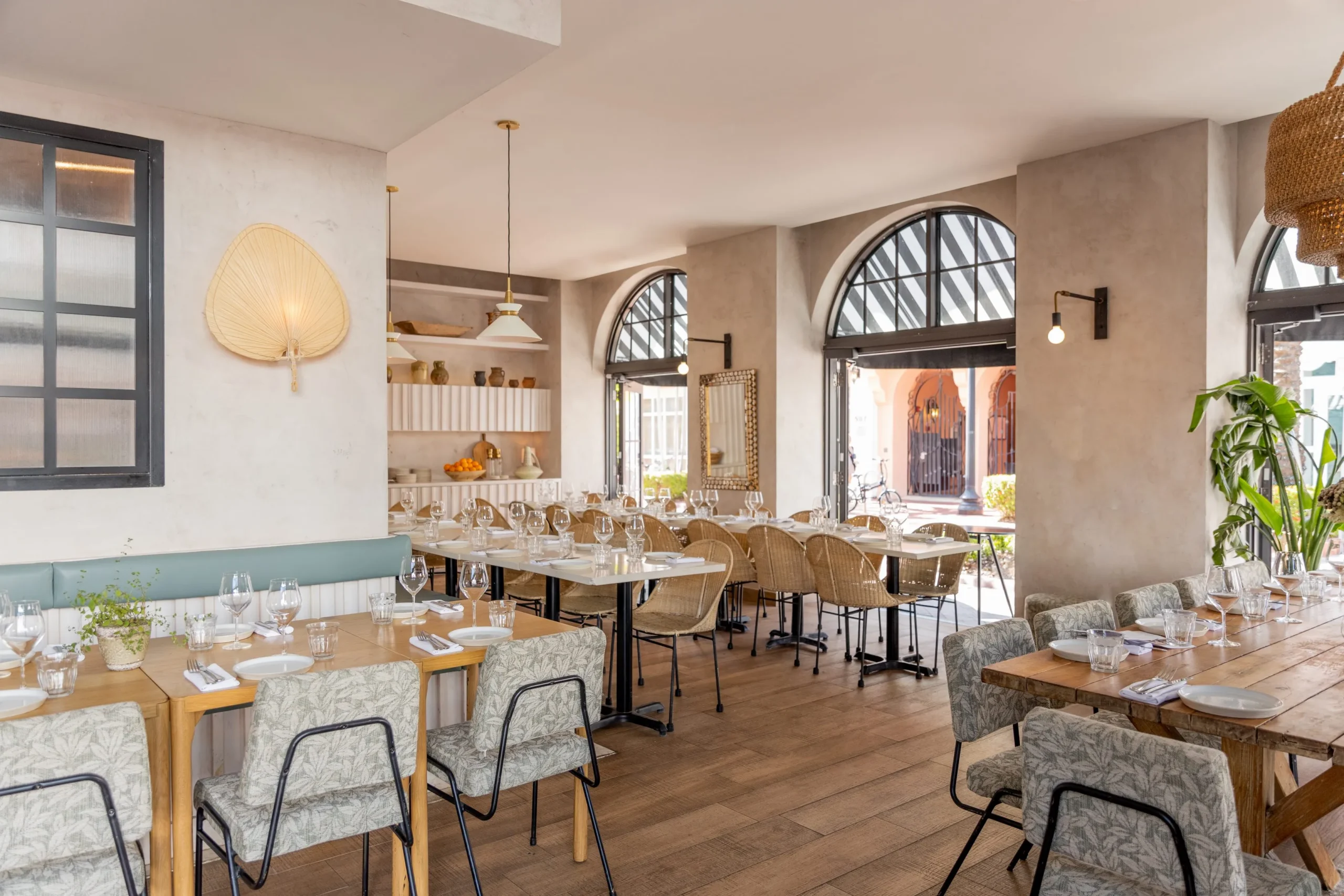 A bright, modern restaurant with wooden floors, light-colored walls, large arched windows, and tables set with glassware and napkins. Woven chairs, decorative accents, and potted plants add a cozy, inviting feel.