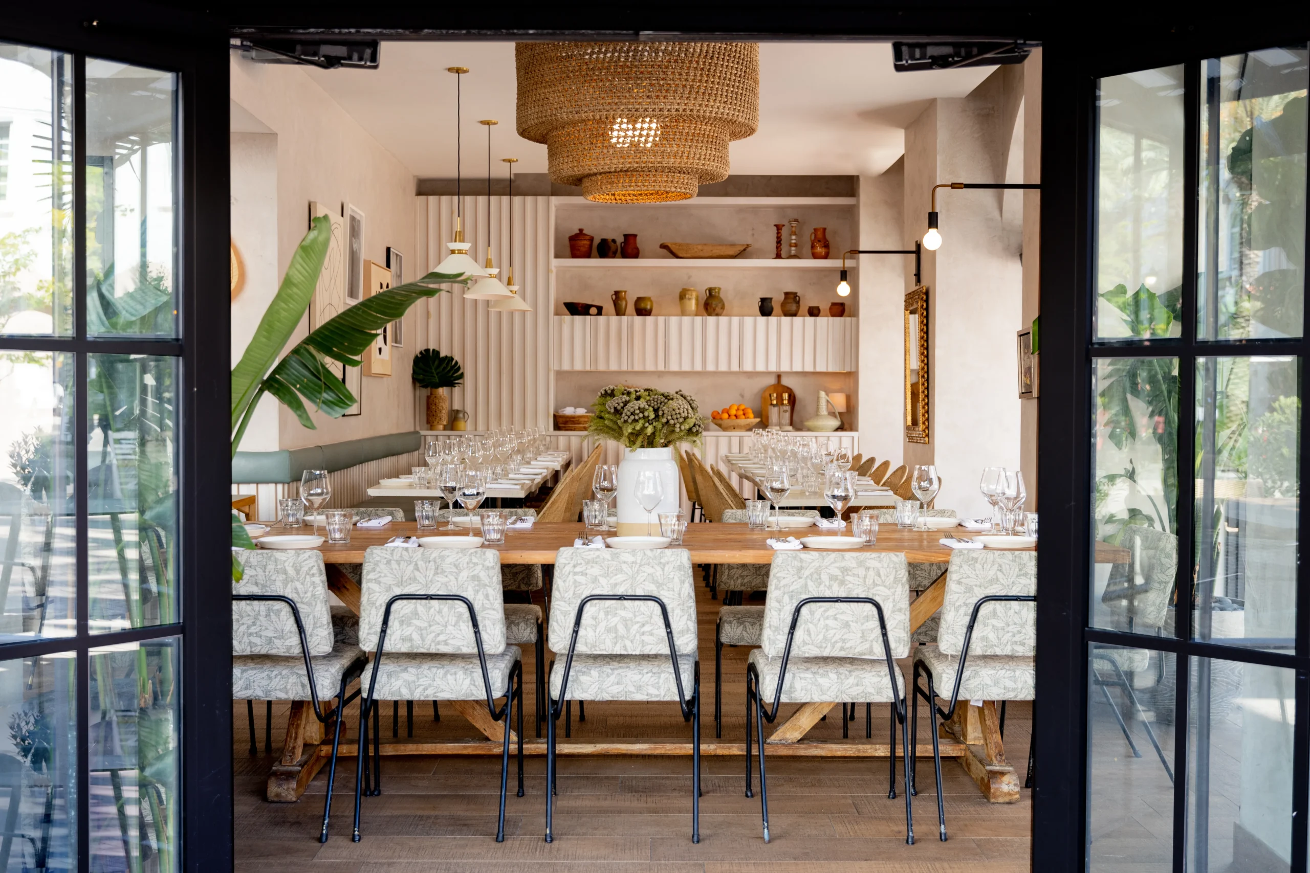 A stylish, modern dining room with a long wooden table set for a meal, cushioned chairs, decorative shelves, large plant, pendant lighting, and abundant natural light shining through glass doors.