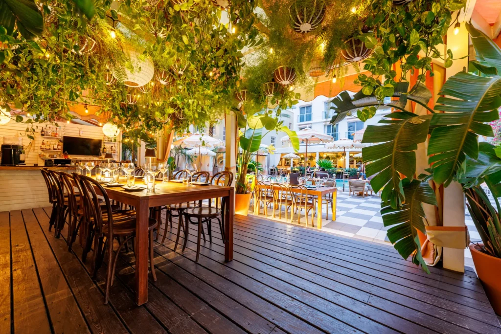 An inviting outdoor restaurant features wooden tables, chairs, string lights, and lush hanging greenery under a pergola, with more seating and umbrellas visible in a sunlit courtyard.