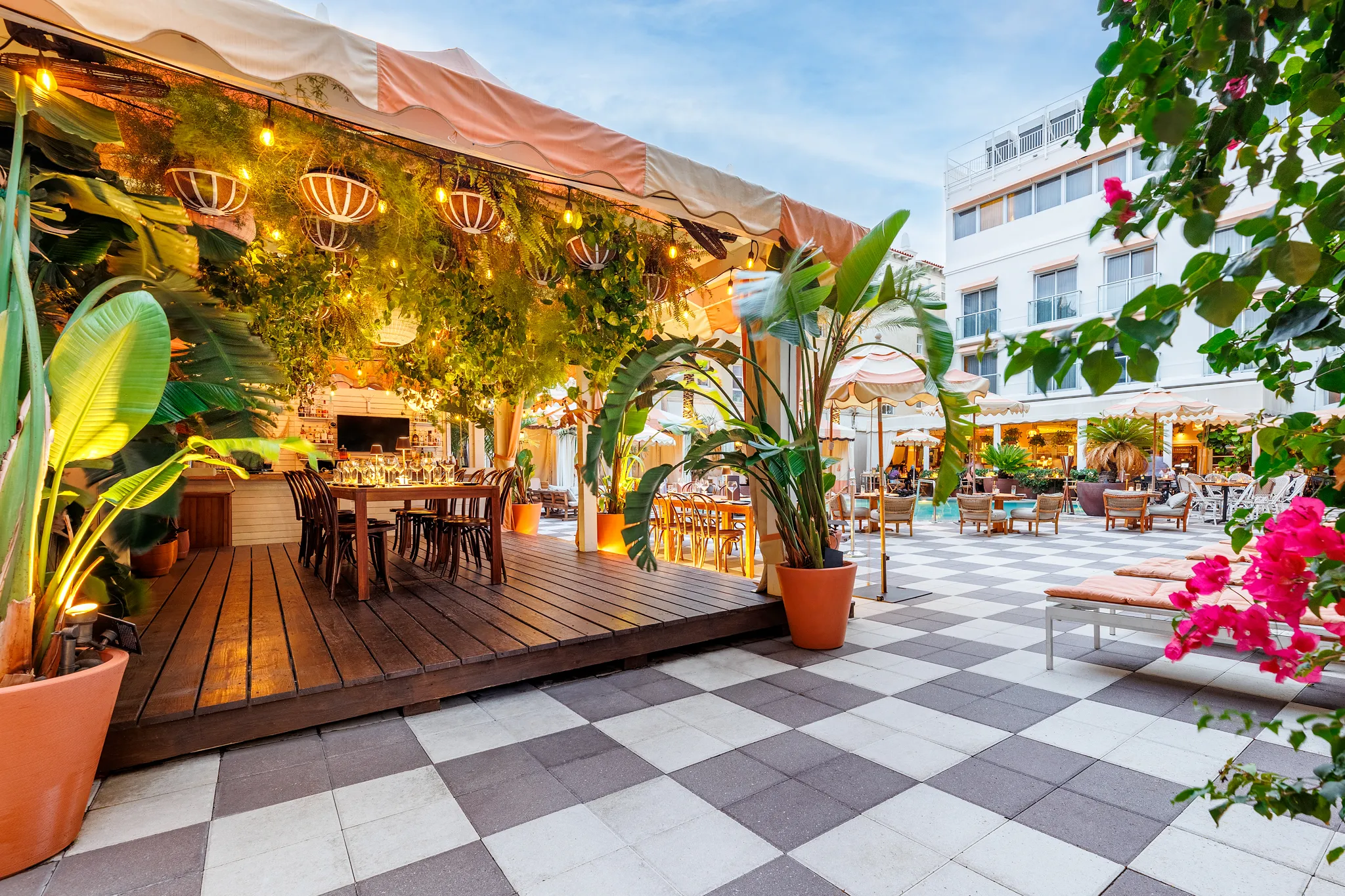 Outdoor restaurant patio with hanging lights, lush green plants, and wooden dining tables under a canopy. The space features checkered tile flooring, potted plants, and nearby lounge seating with a pool in the background.