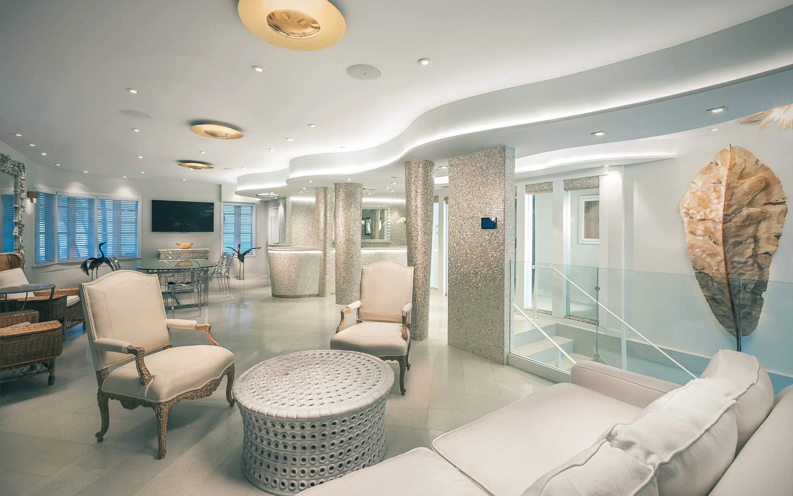 Bright, modern lounge with elegant white and beige furniture, round metal coffee table, textured columns, soft lighting, large wall art of a leaf, and windows with blue shutters.