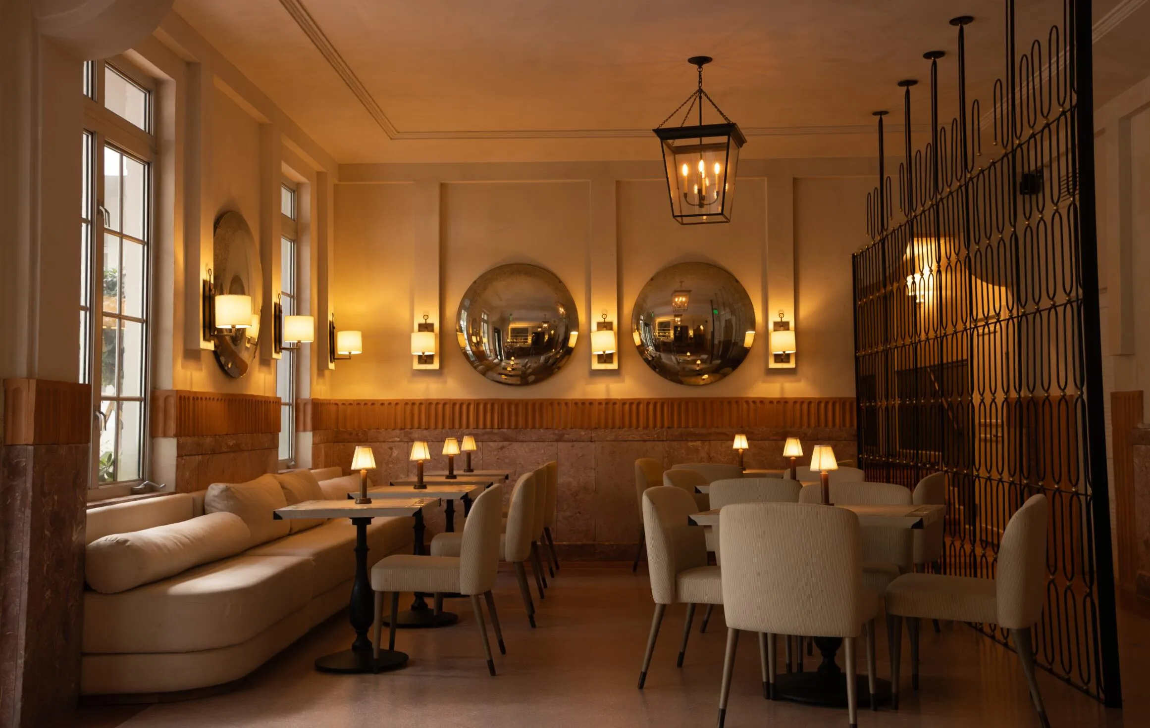 A warmly lit, elegant restaurant dining area with cream-colored chairs, round tables, a cushioned bench, wall sconces, and three large round mirrors reflecting soft light.