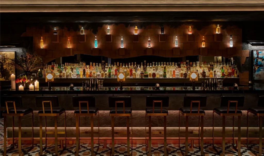 A stylish bar with a row of empty barstools, a patterned tile floor, and shelves filled with various bottles of liquor, softly lit by warm accent lighting and candles.