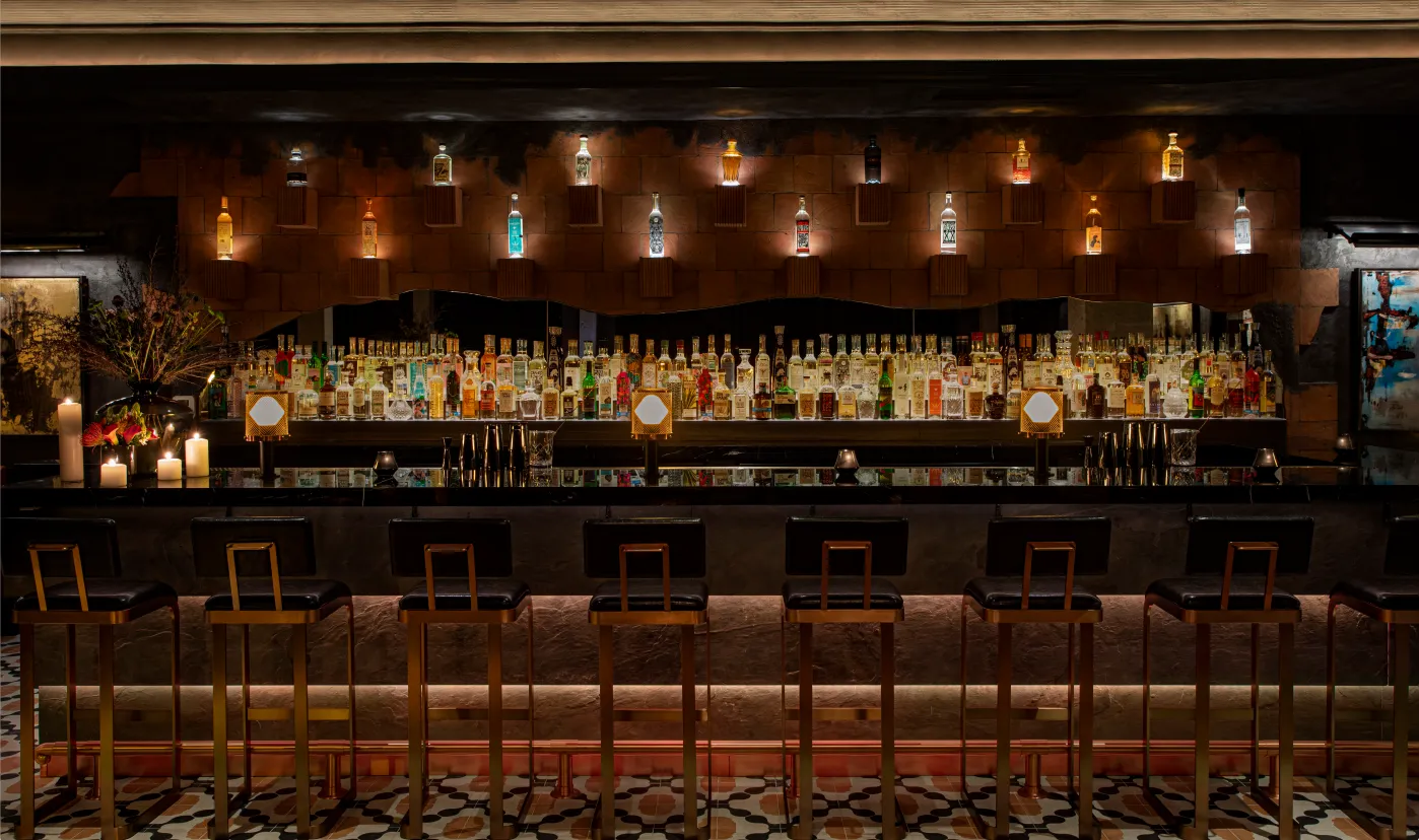 A stylish bar with a row of empty barstools, a patterned tile floor, and shelves filled with various bottles of liquor, softly lit by warm accent lighting and candles.